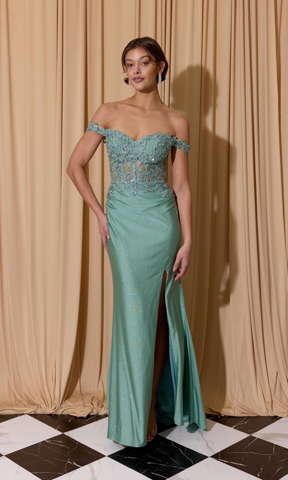 A woman wearing the Eureka EK173 Long Prom Dress, a light green off-the-shoulder gown with lace bodice, thigh-high slit, and floral embroidered corset detail, stands before beige curtains on a black and white tiled floor.